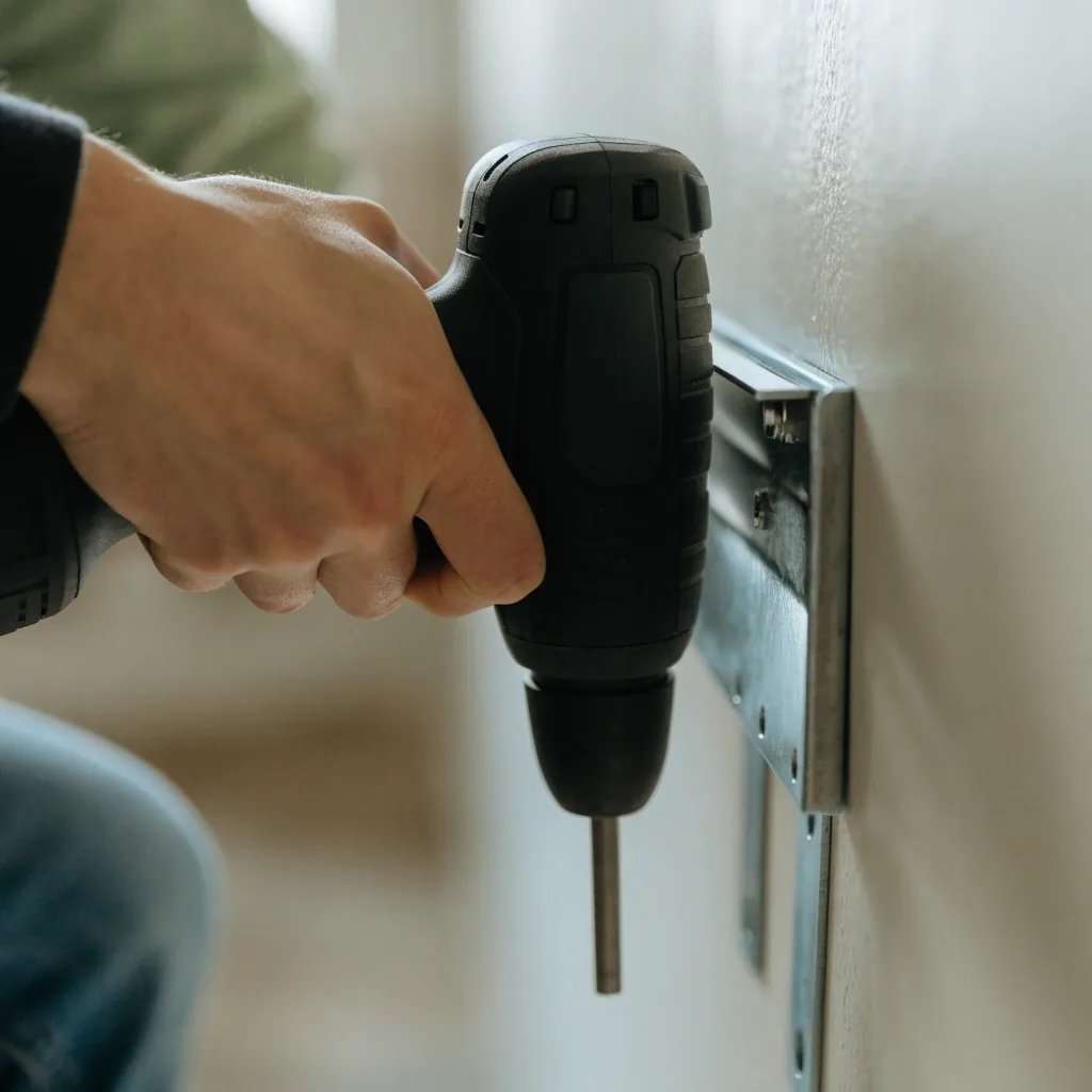 Hands using a cordless drill to mount a shelf bracket on a pale wall